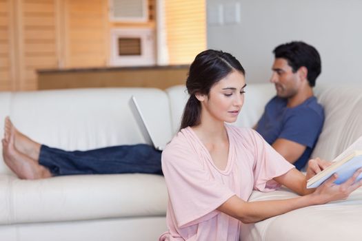 Woman reading a book while her fiance is using a laptop in their living room
