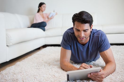 Man using a tablet computer while his girlfriend is reading a book in their living room