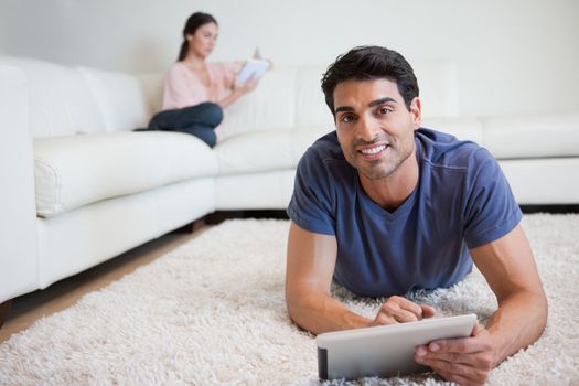 Man using a tablet computer while his fiance is reading a book in their living room