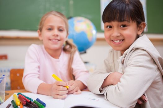 Smiling schoolgirls drawing while looking at the camera in a classroom