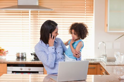 Young mother and daughter with cellphone and notebook in the kitchen together