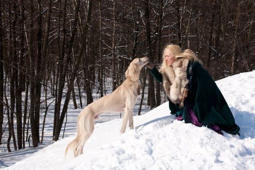 A blonde girl and a standing white saluki on snow
