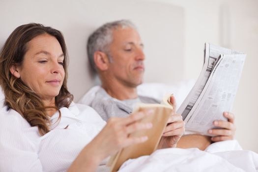 Woman reading a book while her husband is reading the news in their bedroom