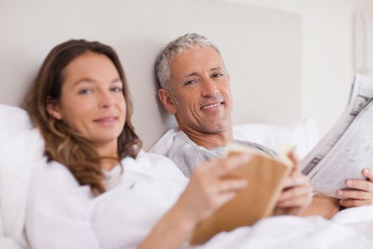 Happy woman reading a book while her husband is reading the news in their bedroom