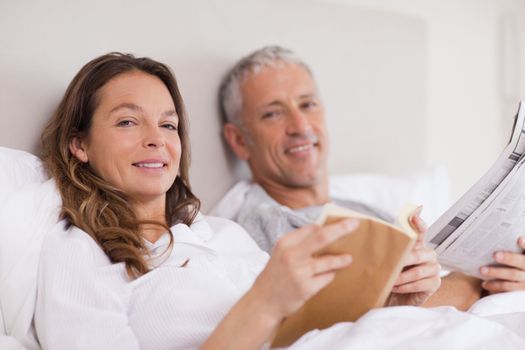 Smiling woman reading a book while her husband is reading the news in their bedroom