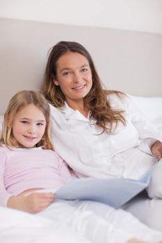 Portrait of a smiling girl reading a book with her mother in a bedroom
