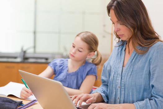Little girl doing her homework while her mother is using notebook in a kitchen