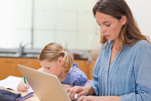 Little girl doing her homework while her mother is using laptop in a kitchen