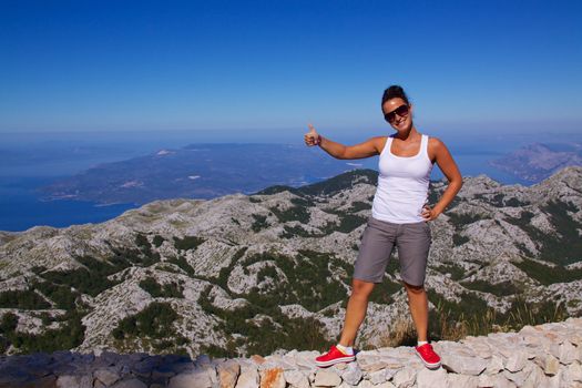 Smiling young girl on hilltop showing thumbs above beautiful panorama of nature park Biokovo on Dalmatian coast near Makarska Riviera in Croatia