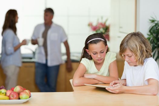 Children using tablet in the kitchen with their parents behind them