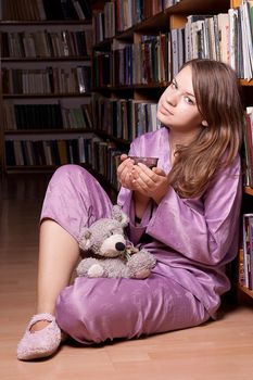 The girl in purple pajamas with a toy sits on a shelf of books in the library of photography