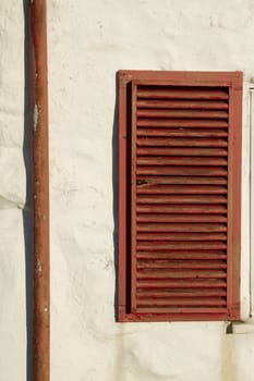 A rustic wooden, brown painted, window shutter and downpipe on a cream painted stone wall.