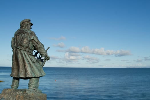 A bronze statue of a sea captain with coat and hat holding a ship's wheel looking out to sea.