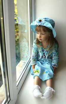 Cute thoughtful  little girl in blue dress looking through the window