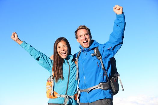 Happy hiking couple cheering in winning success concept. Young woman and man hiker holding around eachother elated and cheerful with arms raised in the sky. Asian girl and Caucasian male hikers.