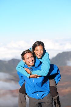 Hiking couple piggybacking happy and cheerful on hike in high mountains. Joyful smiling interracial couple having fun together. Caucasian man hiker and Asian woman hiker.