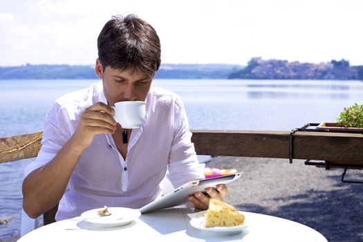 Good looking man drinking cappuccino in front of lake working with electronic tablet