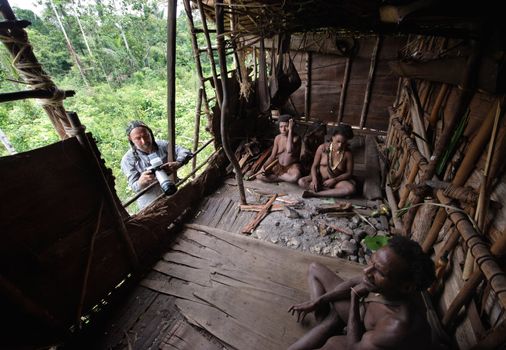 INDONESIA, NEW GUINEA, IRIAN JAYA, ONNI VILLAGE - JUNY 27: White man with camera and Korowai people in the traditional wooden house built on a tree. New Guinea Island, Indonesia. June 27 2012