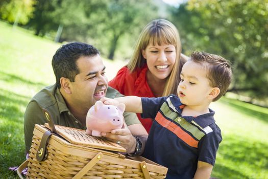 Happy Mixed Race Couple Give Their Son a Piggy Bank at a Picnic in the Park.