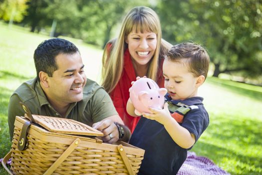 Happy Mixed Race Couple Give Their Son a Piggy Bank at a Picnic in the Park.