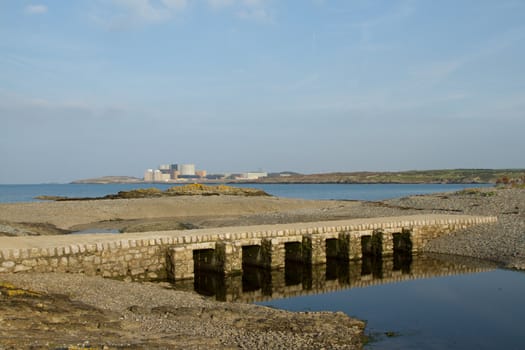 Water leads to a bridge built as a causeway crossing a tidal waterway with pebbles with the sea and an industrial complex in the background.