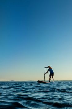 Woman paddling stand up paddle board at sunset with blue sky in background.