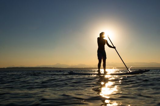 Woman paddling stand up paddle board at sunset with mountains in background.