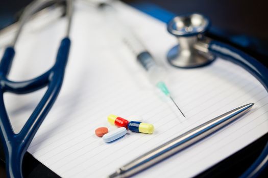Note pad with stethoscope and pen along with  serynge and capsules on a blue and dark background