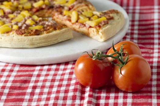 A horizontal image of hawaiian pizza and whole ripe tomato on a checkered table cloth 