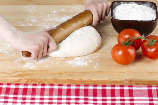 Female hand with rolling pin kneading pizza dough with flour and tomato on the side