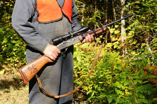 Real man wearing bright orange vest going moose hunting with rifle, carving on handle, in the forest during fall or autumn in northern canada.