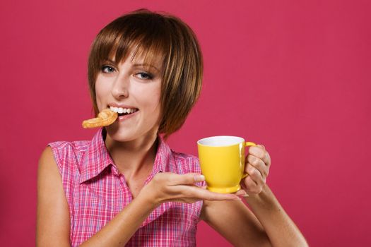 Cute girl with a tea cup biting a pretzel, pink background 
