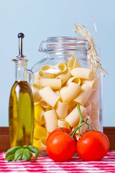 A big glass vase of italian pasta ("Paccheri") near a bottle of oil, tomatoes and basil