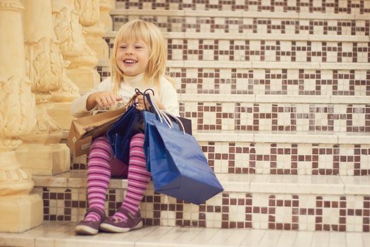 Pleased blonde girl 3 years old in bright clothes sitting on the stairs with shopping