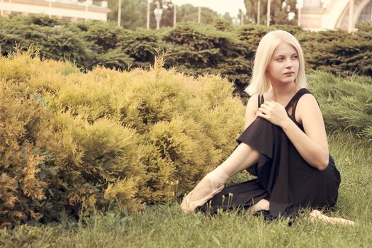 Ballerina in pointe sitting on the grass and looks aside in park