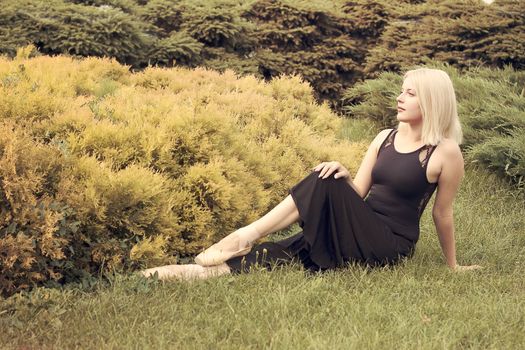 Ballerina in pointe gracefully sitting on the grass and looks forward to the park