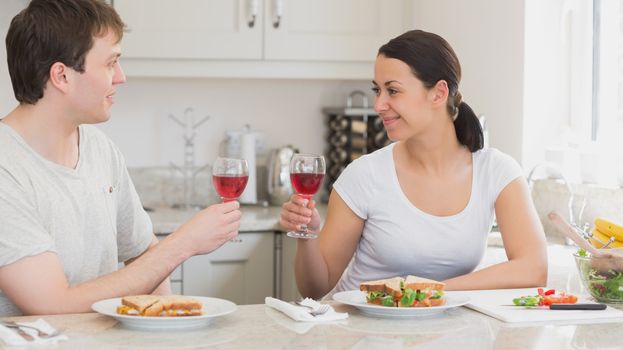 Two people clinking glasses for drinking and eating sandwiches in the kitchen
