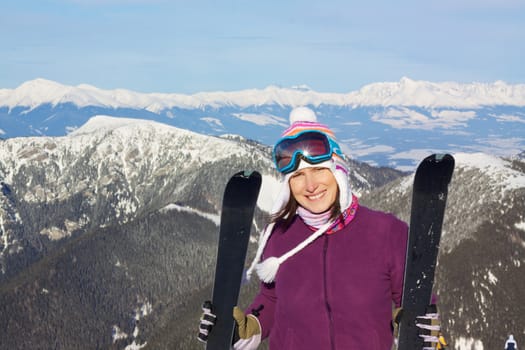 Smiling attractive young girl holding skis with picturesque winter mountainous background