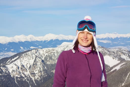 Smiling attractive young woman wearing knitted winter cap and ski glasses with picturesque winter mountains in the background