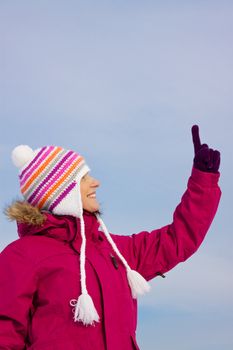 Smiling attractive girl wearing knitted winter cap and gloves pointing upwards with her finger, place for your copy
