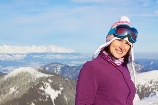 Smiling attractive young woman wearing knitted winter cap and ski glasses with picturesque winter background