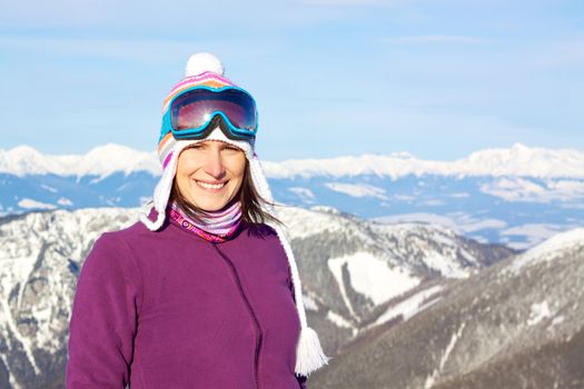Smiling attractive young woman wearing knitted winter cap and ski glasses with picturesque winter background