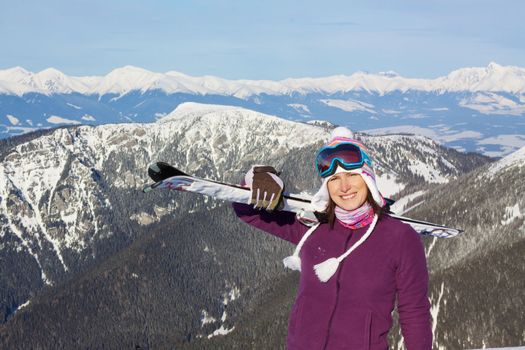 Smiling attractive young girl carrying skis with picturesque winter mountainous background