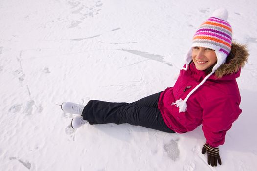 Smiling young woman with ice skates wearing knitted winter cap sitting in snow on frozen lake