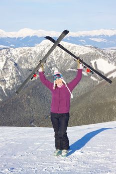 Joyful attractive young girl holding up skis with picturesque winter mountainous background