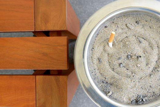 Cigarette in sand ashtray bin is next to wooden bench.