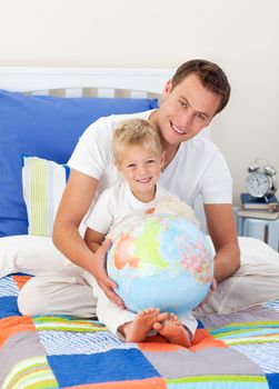 Smiling father and his son looking at a terretrial globe sitting on a bed