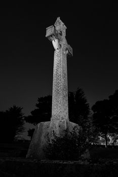 Low angle black and white image of a large stone Celtic cross on a plinth outlined against darkness