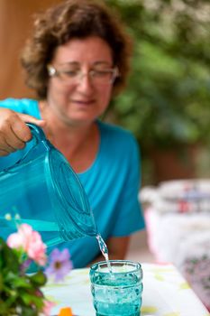 Blue shirt lady is pouring  water in to the blue glass from a blue matching pitcher to drink and cool down. Selective Focus on to action.