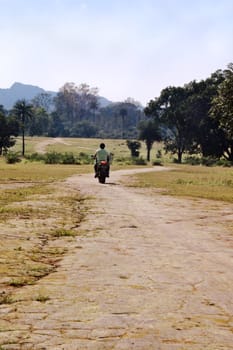 Generic capture of motor biker riding into the horizon over dried mud path through parkland lined with bare grass, trees and bushes. Satura mountains in the distance, location Pachmarhi, Madhya Pradesh, India